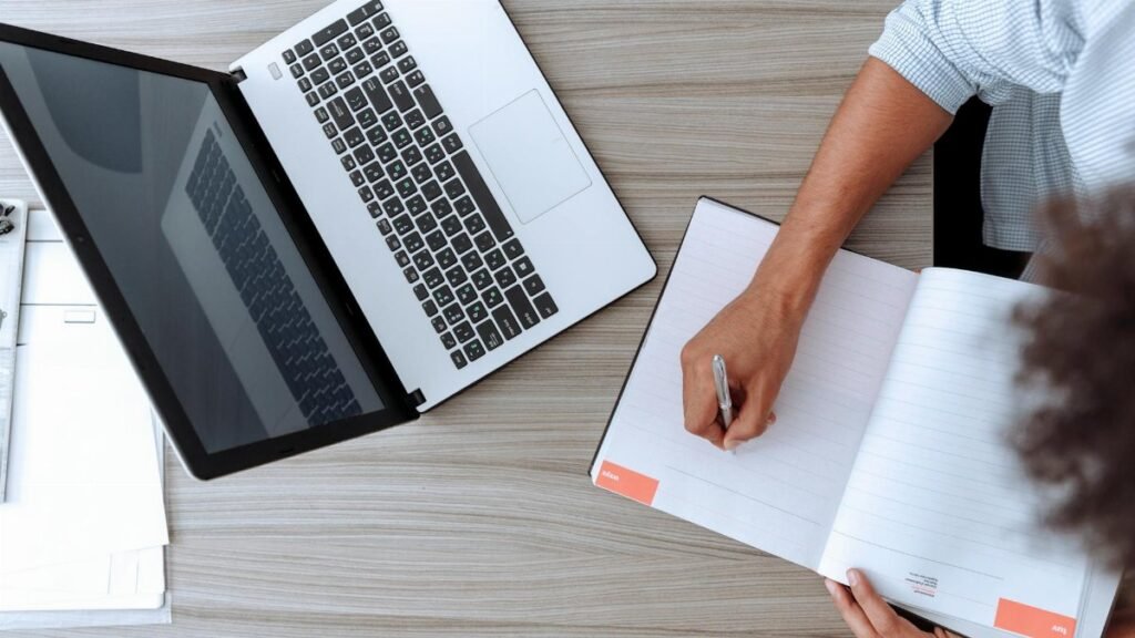 Person writing notes at desk with a laptop and open notebook, capturing a modern workspace.