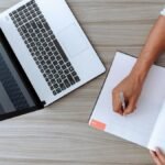 Person writing notes at desk with a laptop and open notebook, capturing a modern workspace.