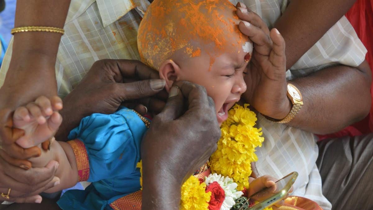 A baby in a traditional Indian ear-piercing ceremony, surrounded by family hands, flowers, and rituals.