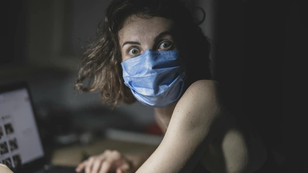 A surprised woman wears a mask and uses a laptop, highlighting pandemic awareness indoors.