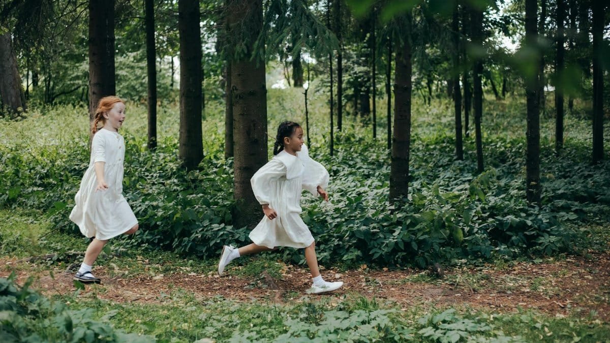 Young girls in white dresses joyfully running through a lush green forest path.