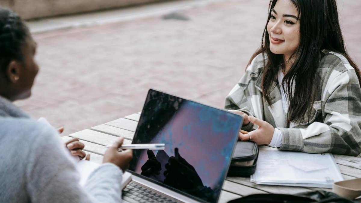 High angle of smiling diverse female students using laptop and taking notes while doing research together at table in campus