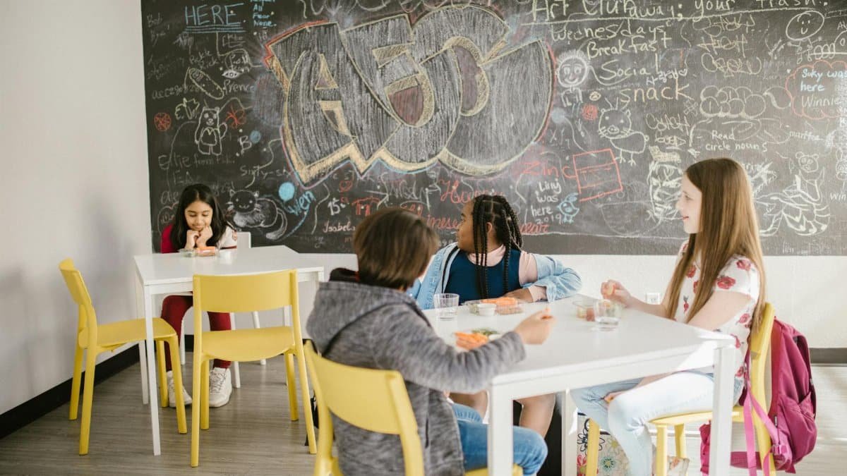 Children sitting in a school classroom having lunch with a chalkboard full of doodles behind them.