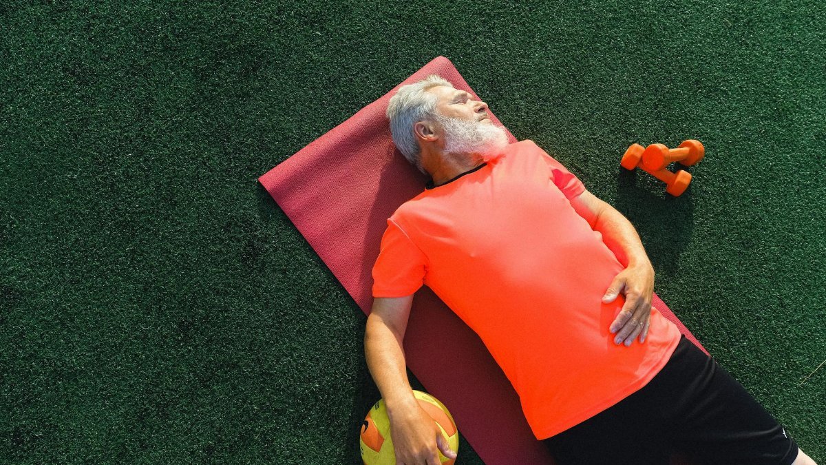 From above of tired senior sportsman lying on fitness mat near ball and dumbbells and resting after training in sunny day