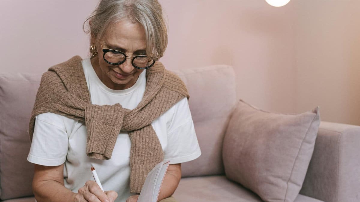 Senior woman with glasses writing in a notebook while sitting on a couch indoors.