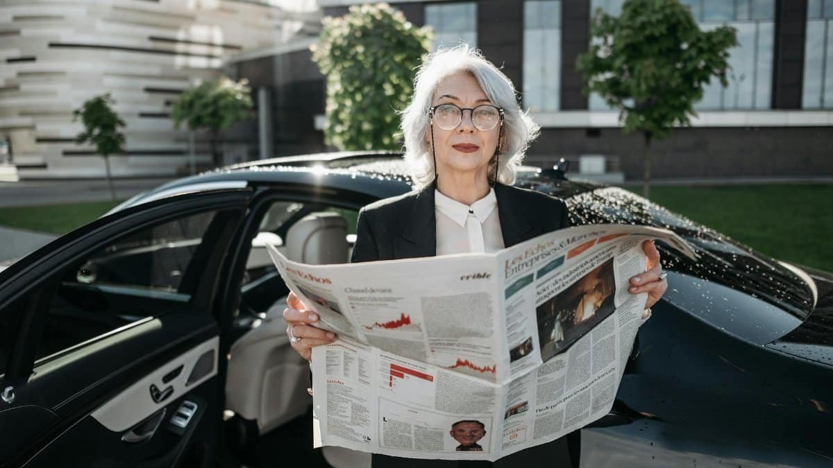 Elderly businesswoman reading a newspaper beside a car outdoors, conveying professionalism and confidence.