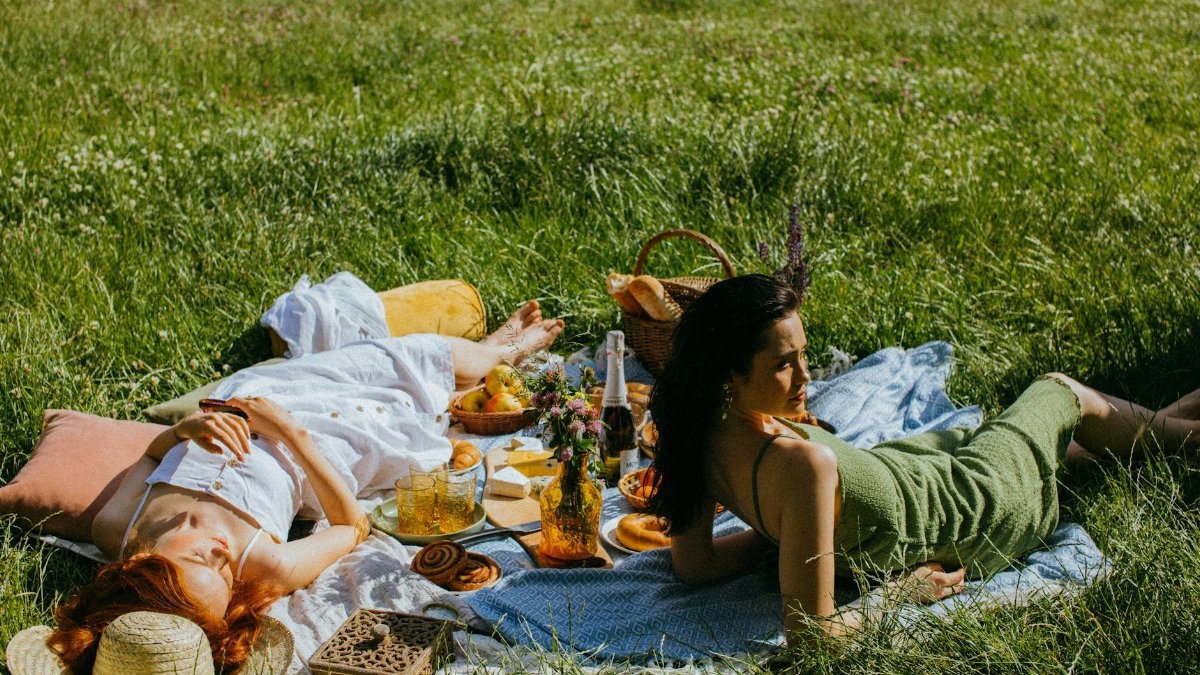 Two women enjoying a relaxing summer picnic on a grass field with fresh food and drinks.