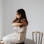 Woman in a light dress sits on wooden table, embracing as a sign of self-care