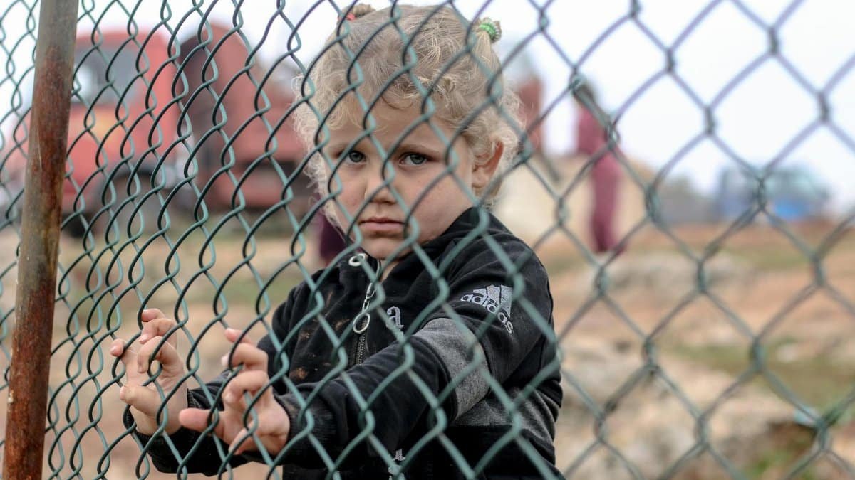A young child stands behind a metal fence in Idlib, Syria, reflecting the challenges faced by refugees.