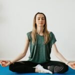 Serene blond lady in sportswear sitting with crossed legs and closed eyes while practicing yoga at home near potted plants