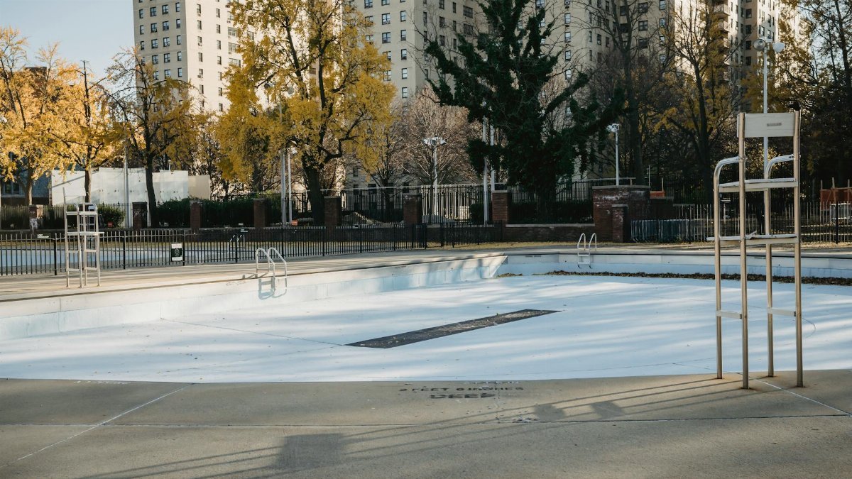 Deserted pool surrounded by high-rise buildings and autumn trees casting shadows in an urban setting.