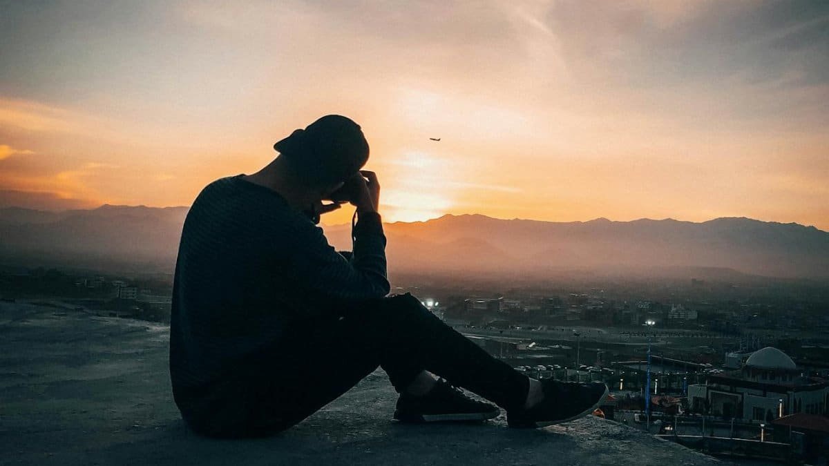 A contemplative silhouette of a man sitting on a rooftop overlooking a city at sunset.