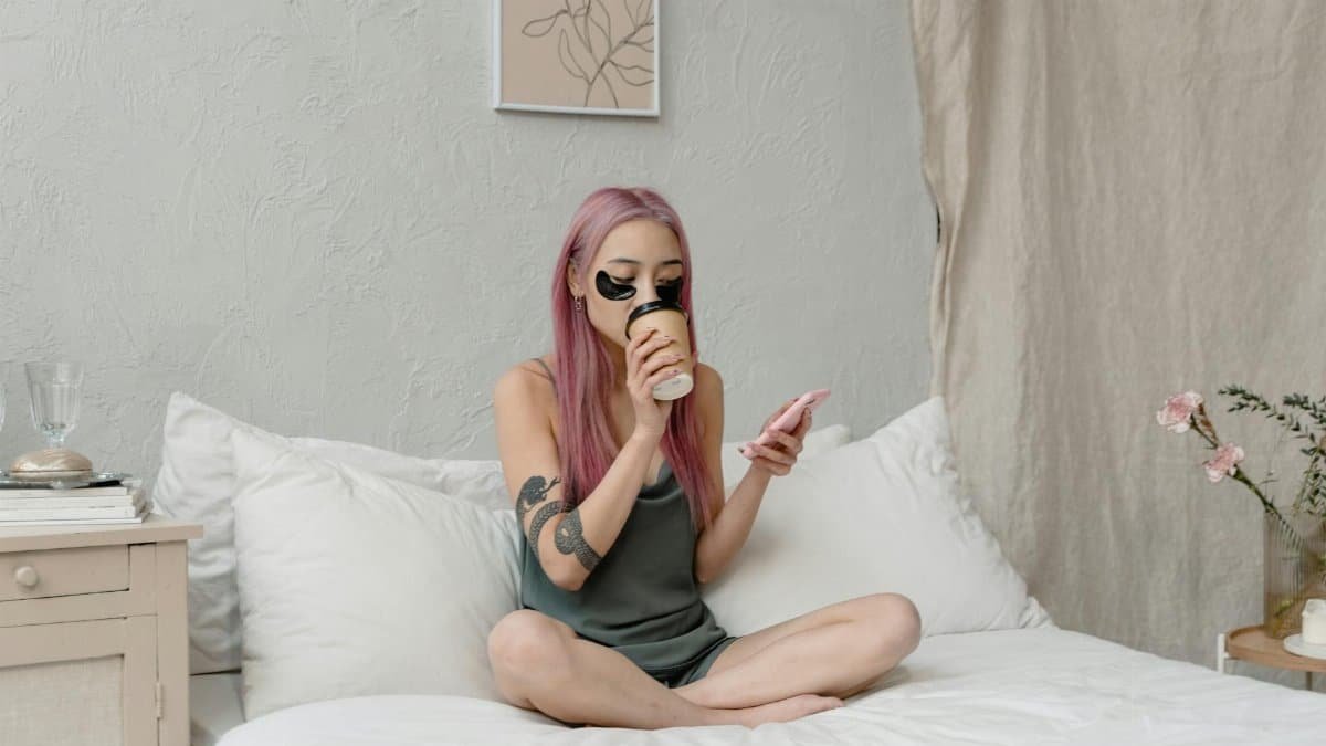 Asian woman enjoys self-care on bed, drinking coffee with under eye patches and phone.
