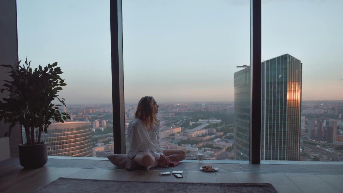 A woman sits by a large window, enjoying the sunset view of skyscrapers in a modern city.