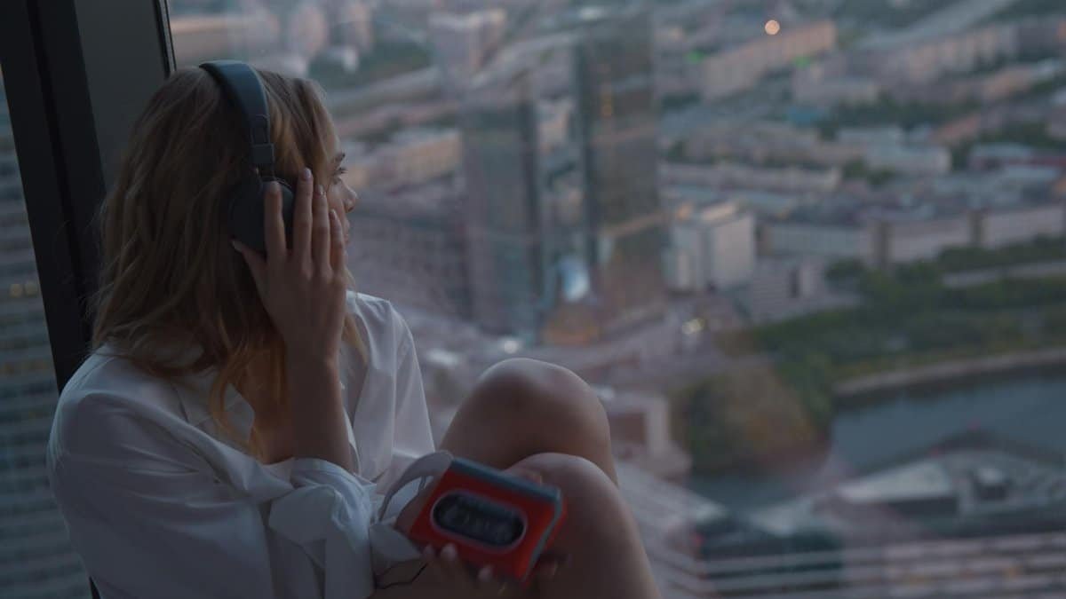 Caucasian woman listening to music on a cassette player by a high-rise window at dusk.