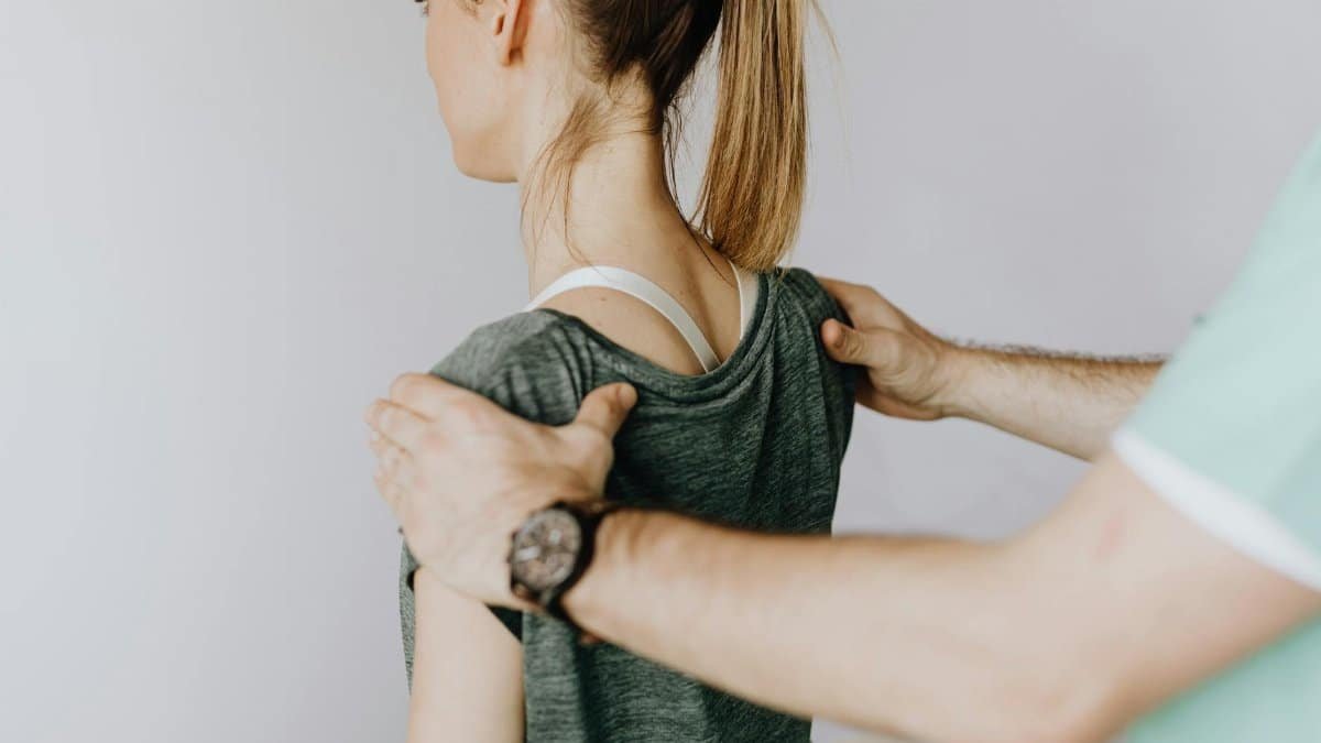 Back view of crop faceless orthopedist in uniform and wristwatch examining shoulder joints of slim female patient in clinic on white background