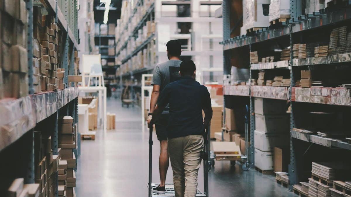Two men maneuver a trolley in a large warehouse filled with boxes and shelves.
