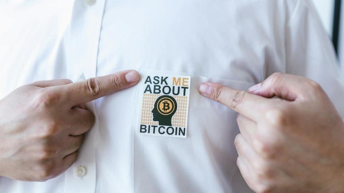 Close-up of a person pointing at a Bitcoin awareness sticker on a shirt.