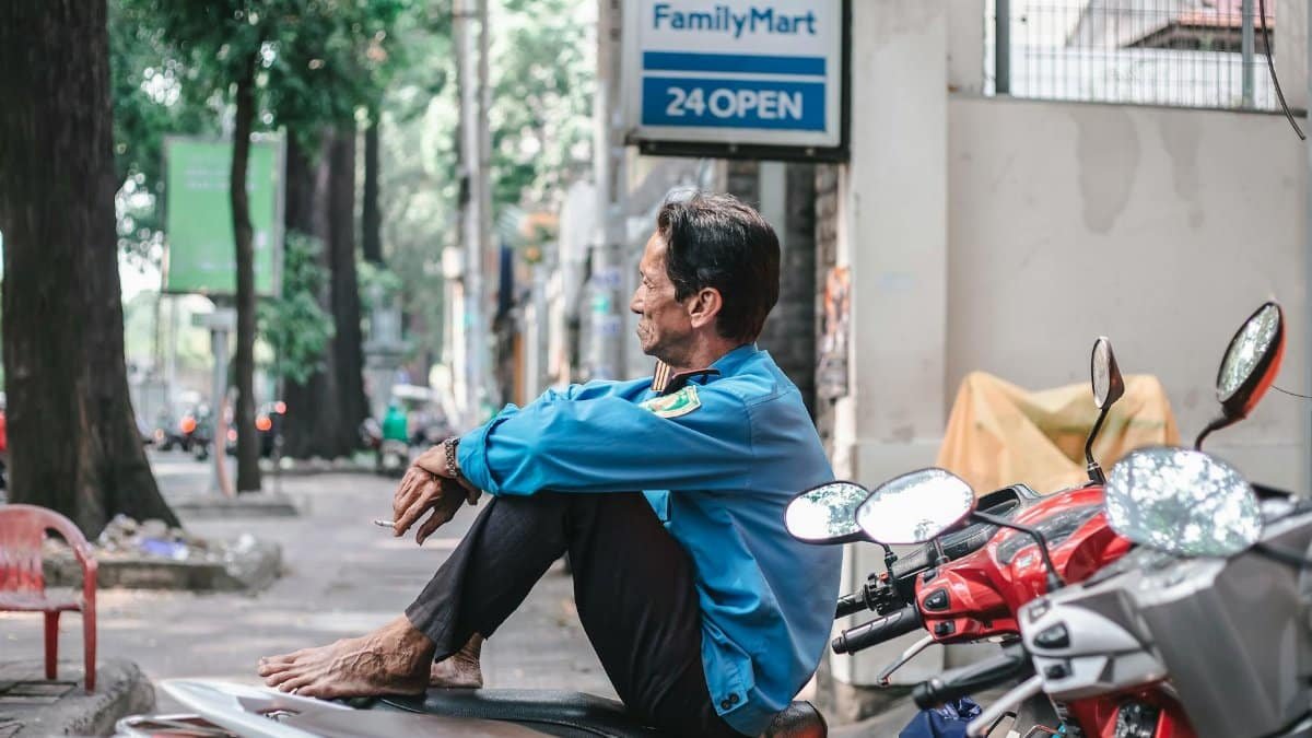 An older man relaxes barefoot on a motorcycle in an Asian city, evoking a moment of calm.