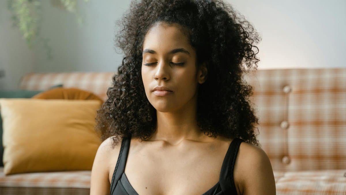 A woman with curly hair meditating indoors with eyes closed. Calm and serene setting.