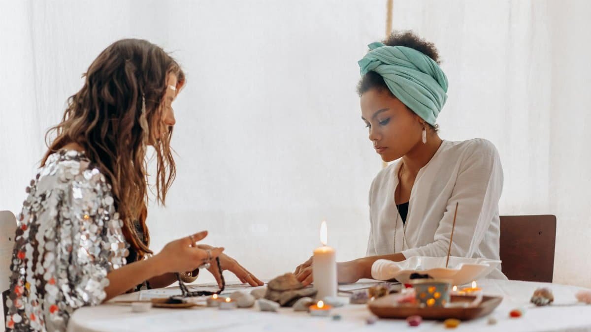 Two women participating in a spiritual ritual with candles and crystals on a table indoors.