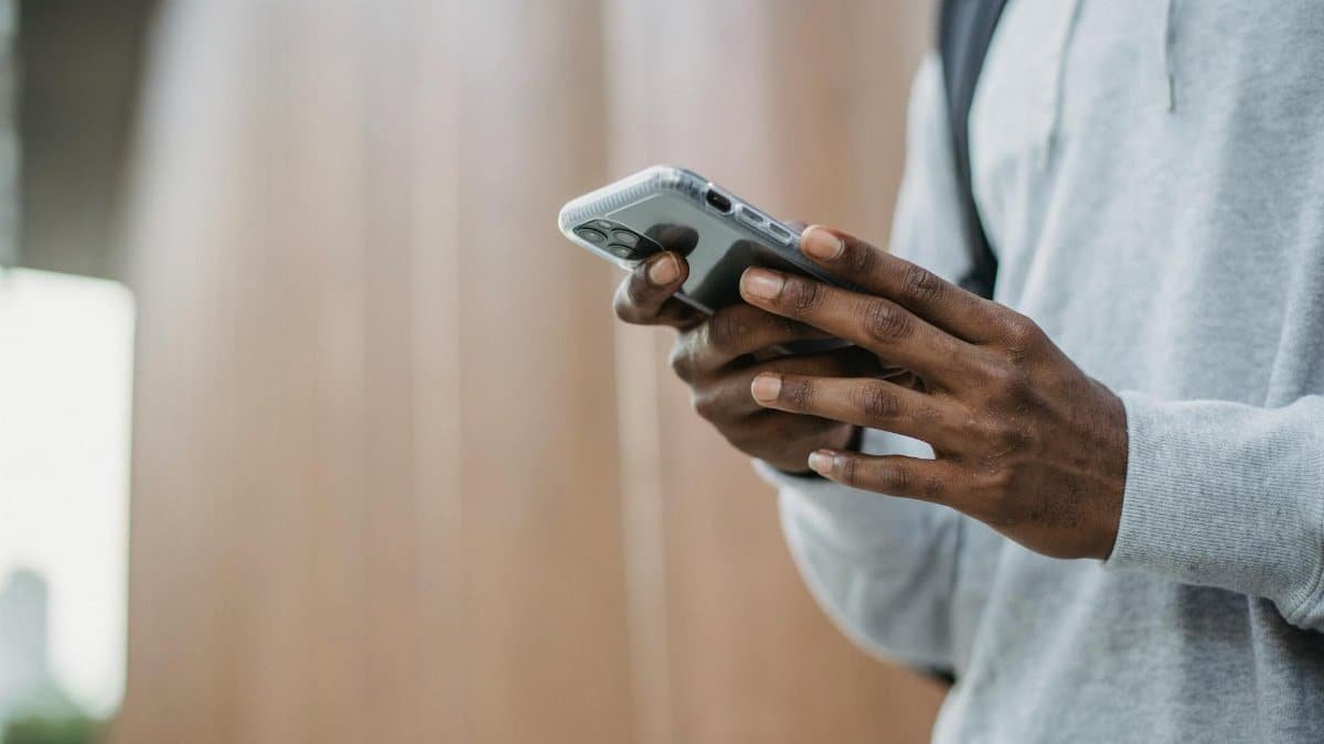 From below crop of unrecognizable African American young guy in casual clothes messaging on mobile phone while standing near wooden wall