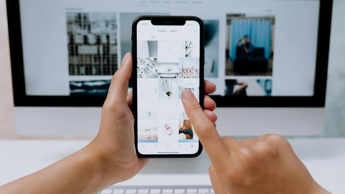 Hands holding a smartphone while browsing a social media app in front of a desktop computer.