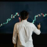 Adult male reviewing stock market data on a large display screen indoors.