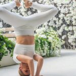 A woman performs a yoga headstand pose indoors surrounded by white flowers, symbolizing balance and fitness.