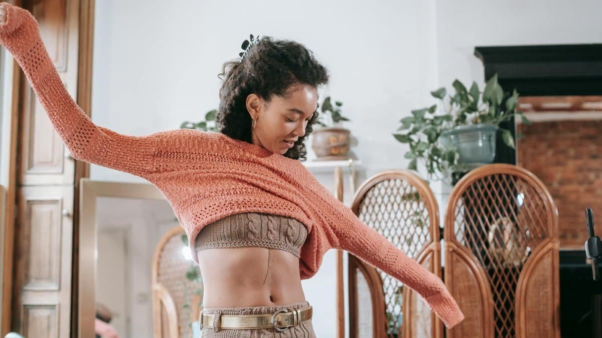 Positive African American female with dark curly hair wearing beige knitted top and leggings with golden belt putting on light orange sweater