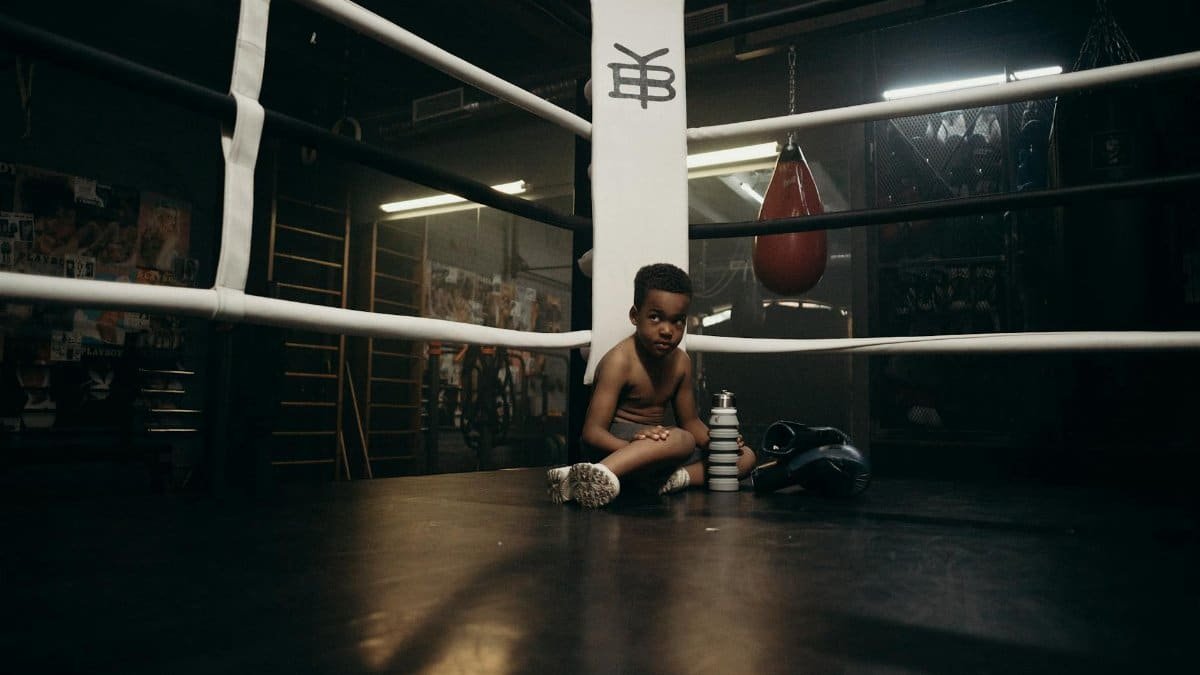A young boy sits in a gym boxing ring, surrounded by training equipment.