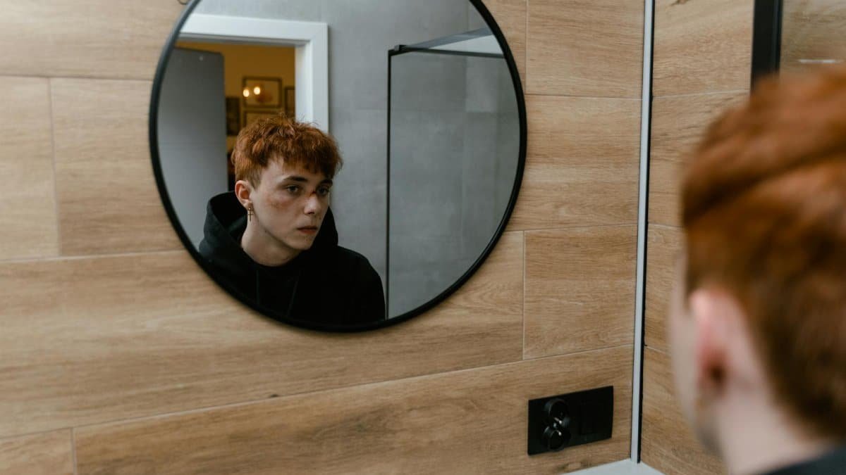 A redheaded teen boy gazes introspectively into a round mirror, reflecting a thoughtful moment.