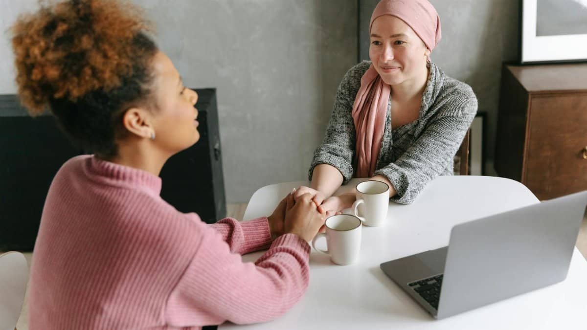 Two women sharing a supportive conversation over coffee indoors. One woman wears a head wrap.