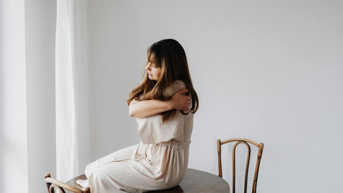 Woman in a light dress sits on wooden table, embracing as a sign of self-care
