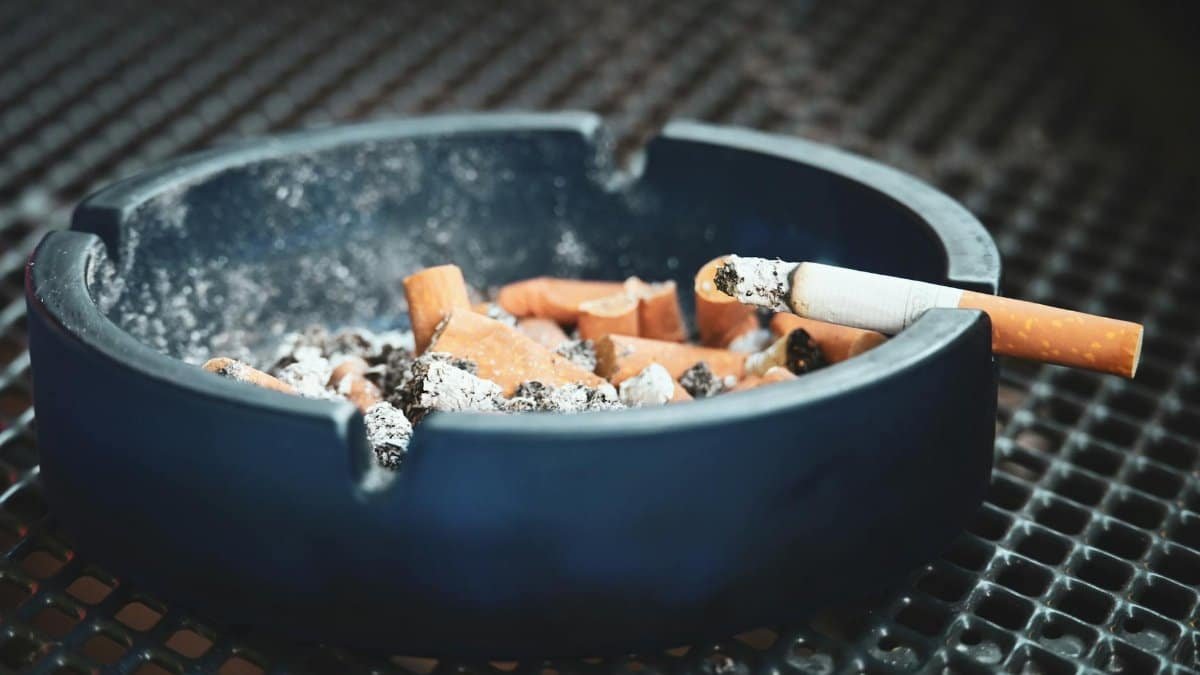 Close-up of cigarette butts and ashes in a blue ashtray with a smoldering cigarette, highlighting smoking habits.