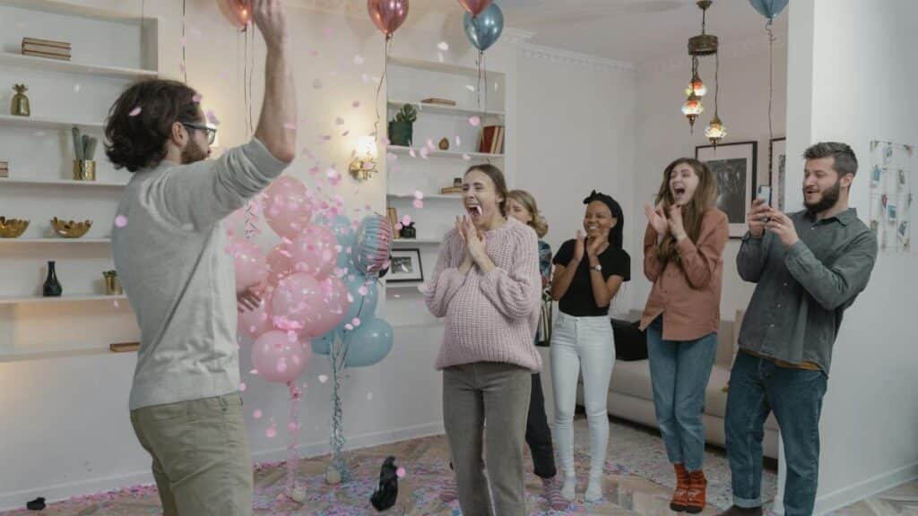 A group of excited people celebrating a gender reveal with balloons and confetti indoors.