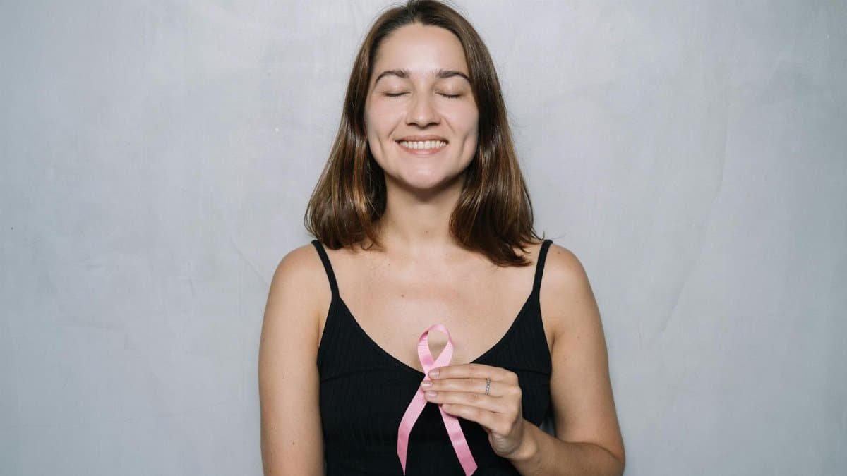 A smiling woman with eyes closed holds a pink breast cancer awareness ribbon, symbolizing hope and health.