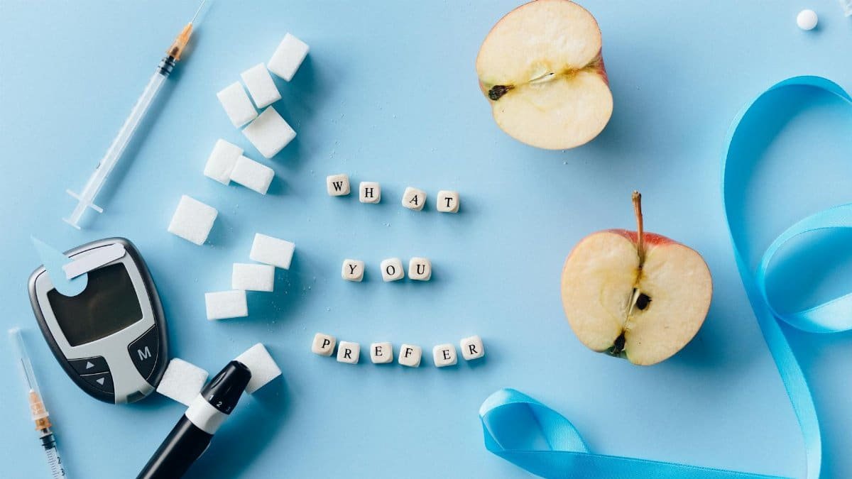Flat lay of diabetes awareness theme with glucometer, syringe, sugar, and apple on blue background.