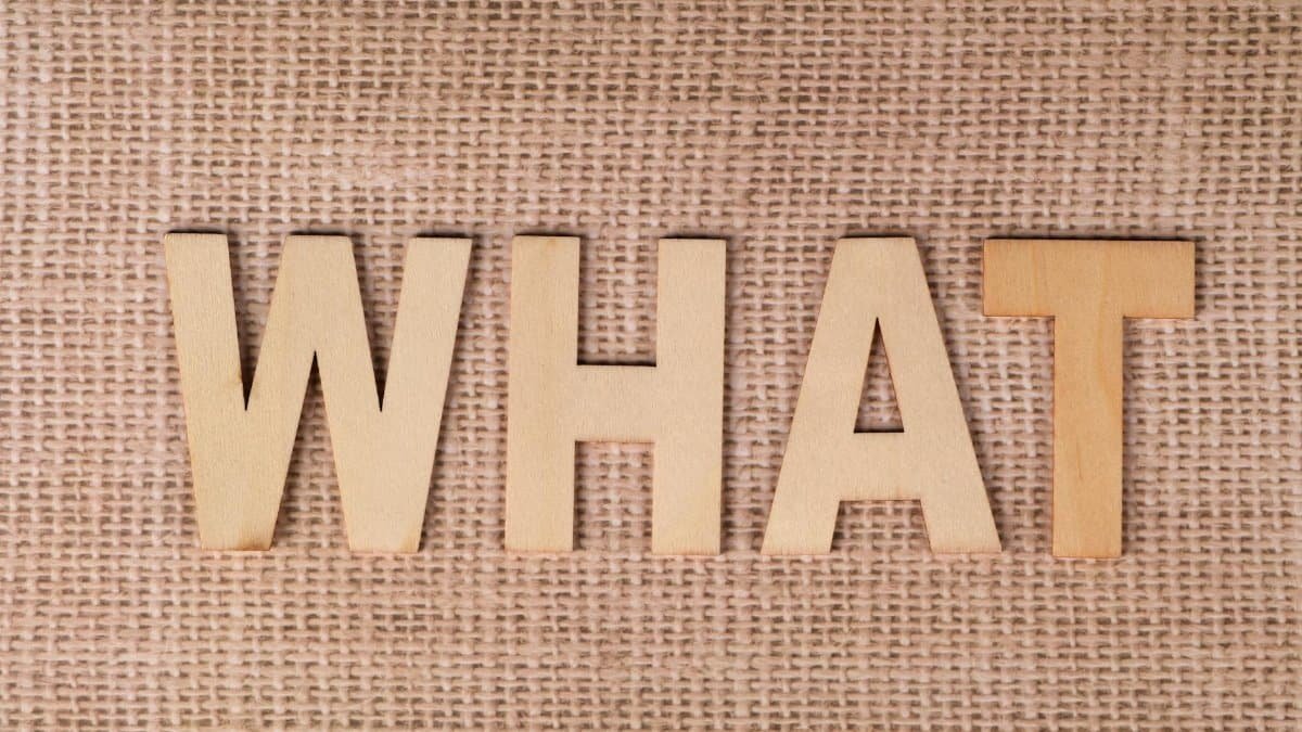Wooden letters forming the word WHAT set on a textured burlap surface.