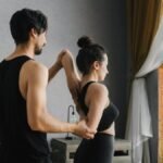 A man and woman practicing partner yoga poses near a large window inside a cozy room.