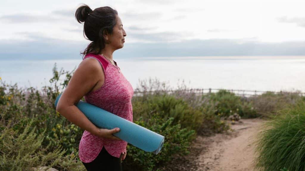 A woman holding a yoga mat stands by the sea, promoting fitness and a healthy lifestyle.