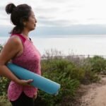 A woman holding a yoga mat stands by the sea, promoting fitness and a healthy lifestyle.