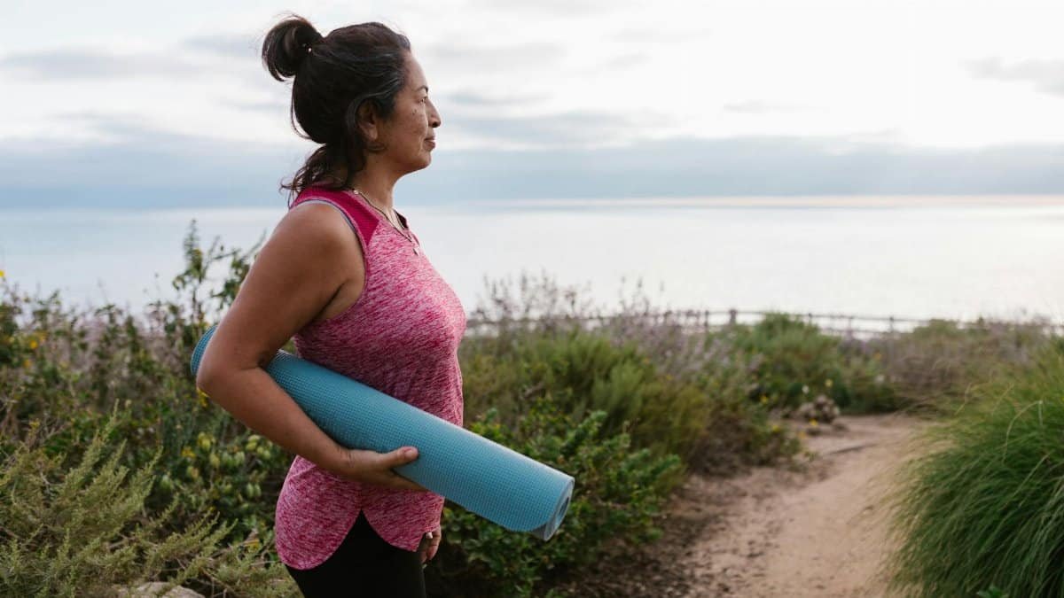 A woman holding a yoga mat stands by the sea, promoting fitness and a healthy lifestyle.