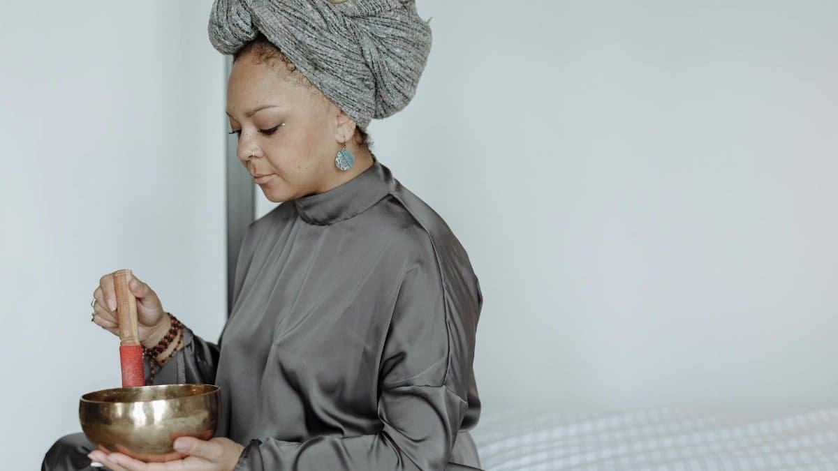 Woman in gray attire meditating indoors with a singing bowl for relaxation.