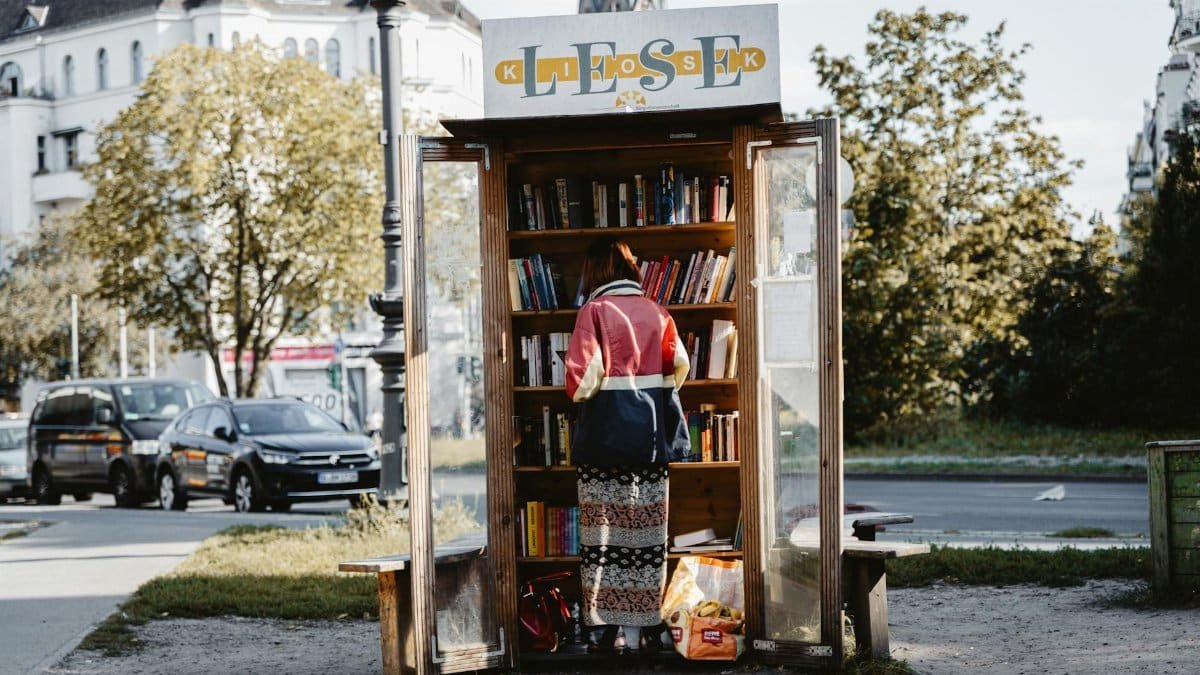 Person using a free library stand on a sunny day with cars and buildings nearby.
