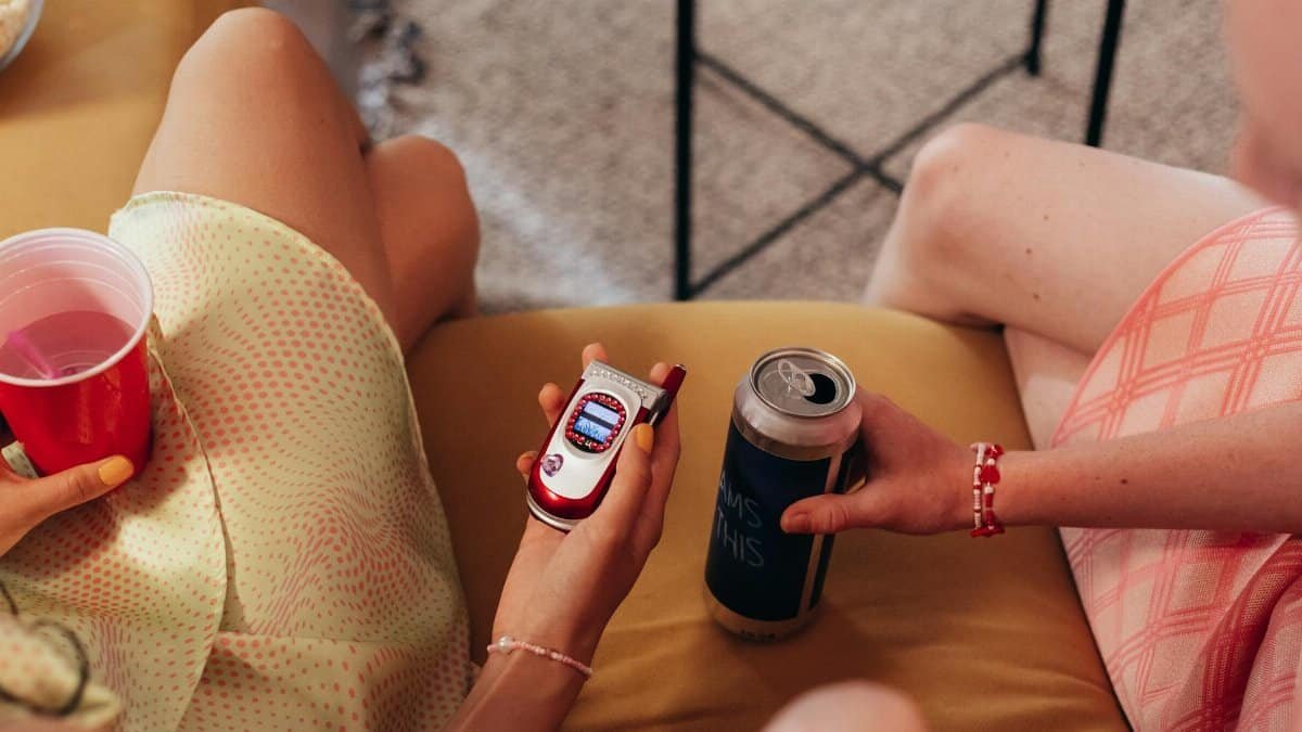 Two women holding drinks and a retro phone sitting indoors, conveying a casual and vintage vibe.