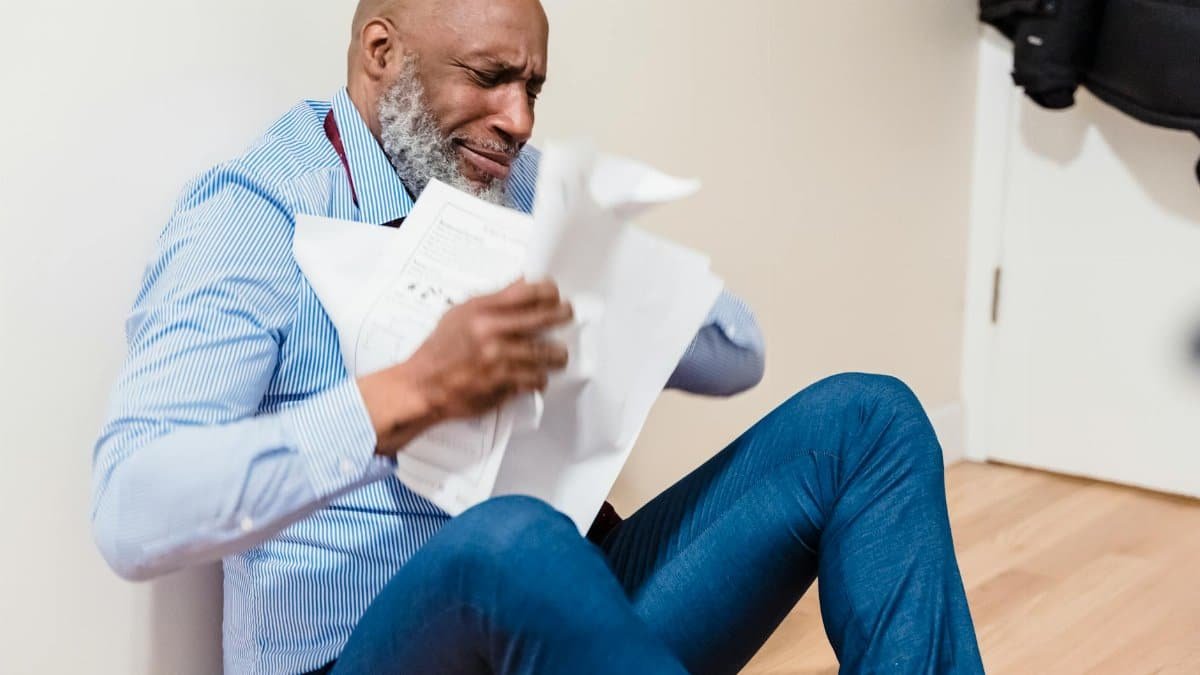 An adult man sitting on the floor, stressed and tearing apart paperwork in frustration.