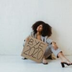 A woman sitting against a wall holding a 'Bye 2020' sign, conveying relief and exhaustion.