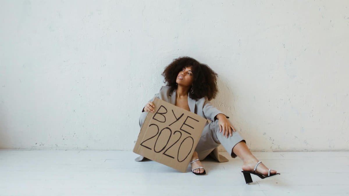 A woman sitting against a wall holding a 'Bye 2020' sign, conveying relief and exhaustion.