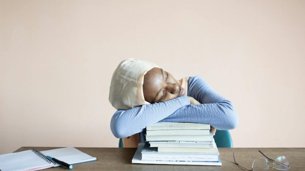 Sleeping African American female student in headscarf sitting at table with opened notebook and lying on stack of textbooks